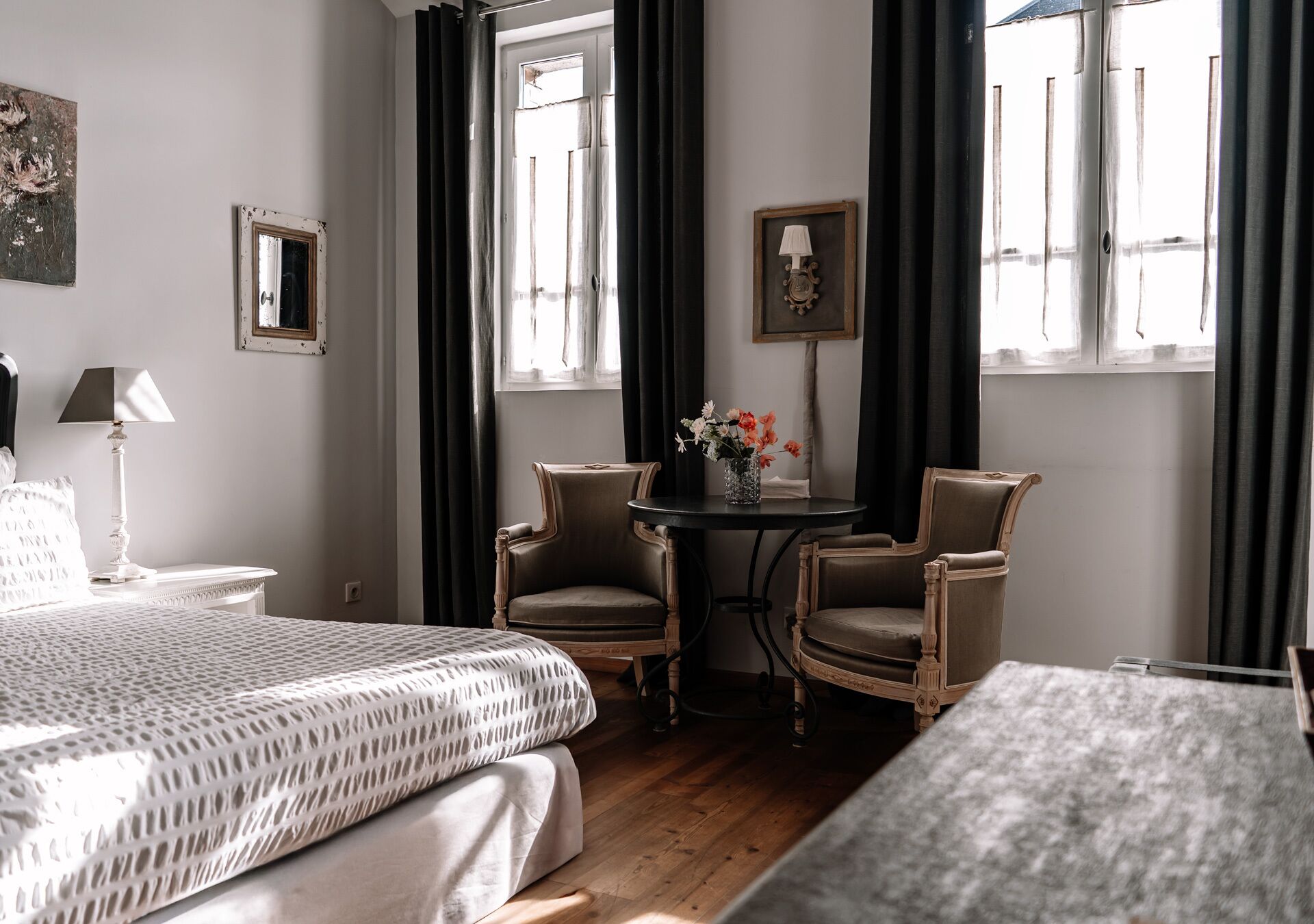 A hotel room with a bed, two armchairs, a small table with a vase of flowers, a lamp, and a window with curtains.