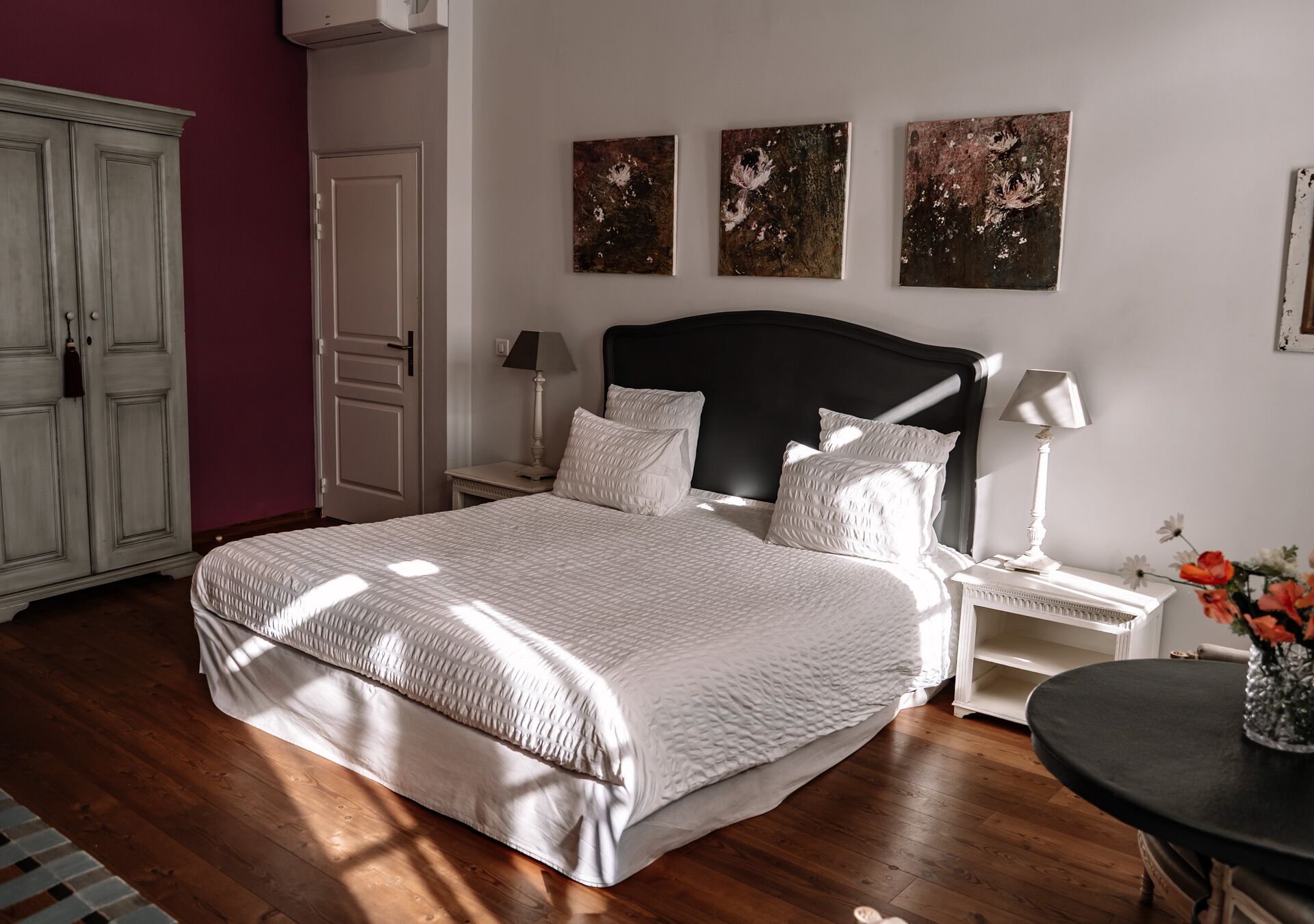 Une chambre à coucher avec un grand lit, du linge de lit blanc, une tête de lit sombre et trois tableaux accrochés au mur.