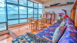 A spacious room with a large window overlooking a body of water, a wooden ceiling fan, a sofa with patterned cushions, and a small table with chairs. - Beautiful condo with ocean view, shared pools and amazing views! (Akumal)