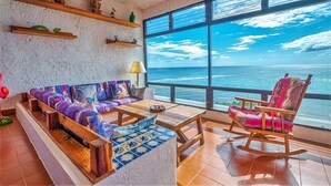A spacious room with a large window offering a view of the ocean. There is a colorful sofa, a rocking chair, and a small table with a bowl of fruit. - Beautiful condo with ocean view, shared pools and amazing views! (Akumal)