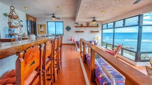 A spacious room with a long wooden bench, a dining table, and chairs. There are ceiling fans, a large window with a view of the ocean, and shelves with decorative items. - Beautiful condo with ocean view, shared pools and amazing views! (Akumal)