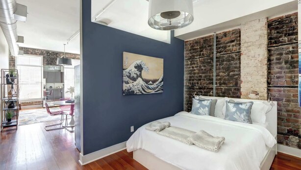 A bedroom with a large bed, a hanging light fixture, a wall-mounted artwork, and a brick wall. - The Loft on Broughton, Modern | Stylish | Chic (Savannah)