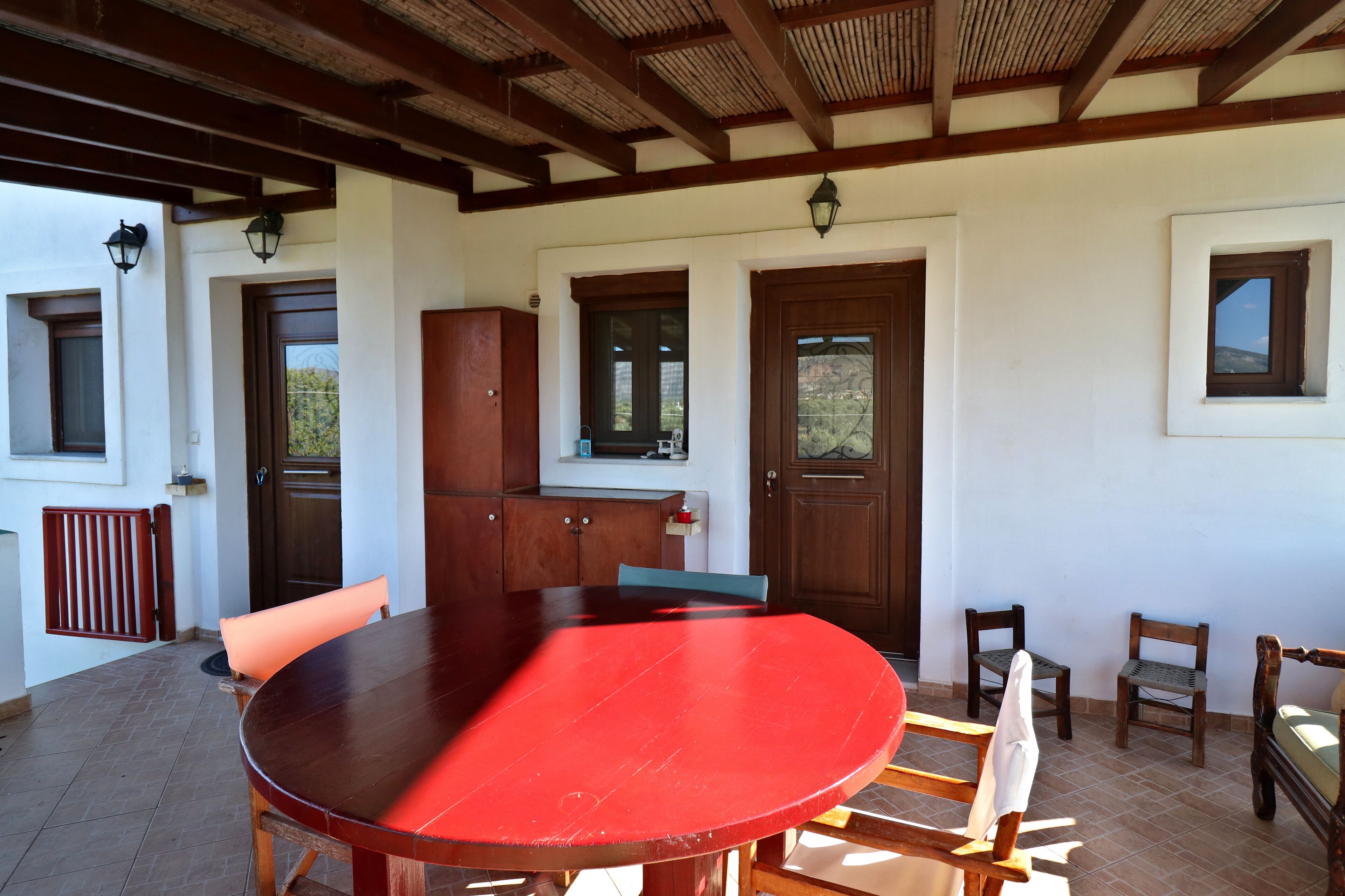 A covered patio area with a red table, wooden chairs, a cabinet, and a door with a window.