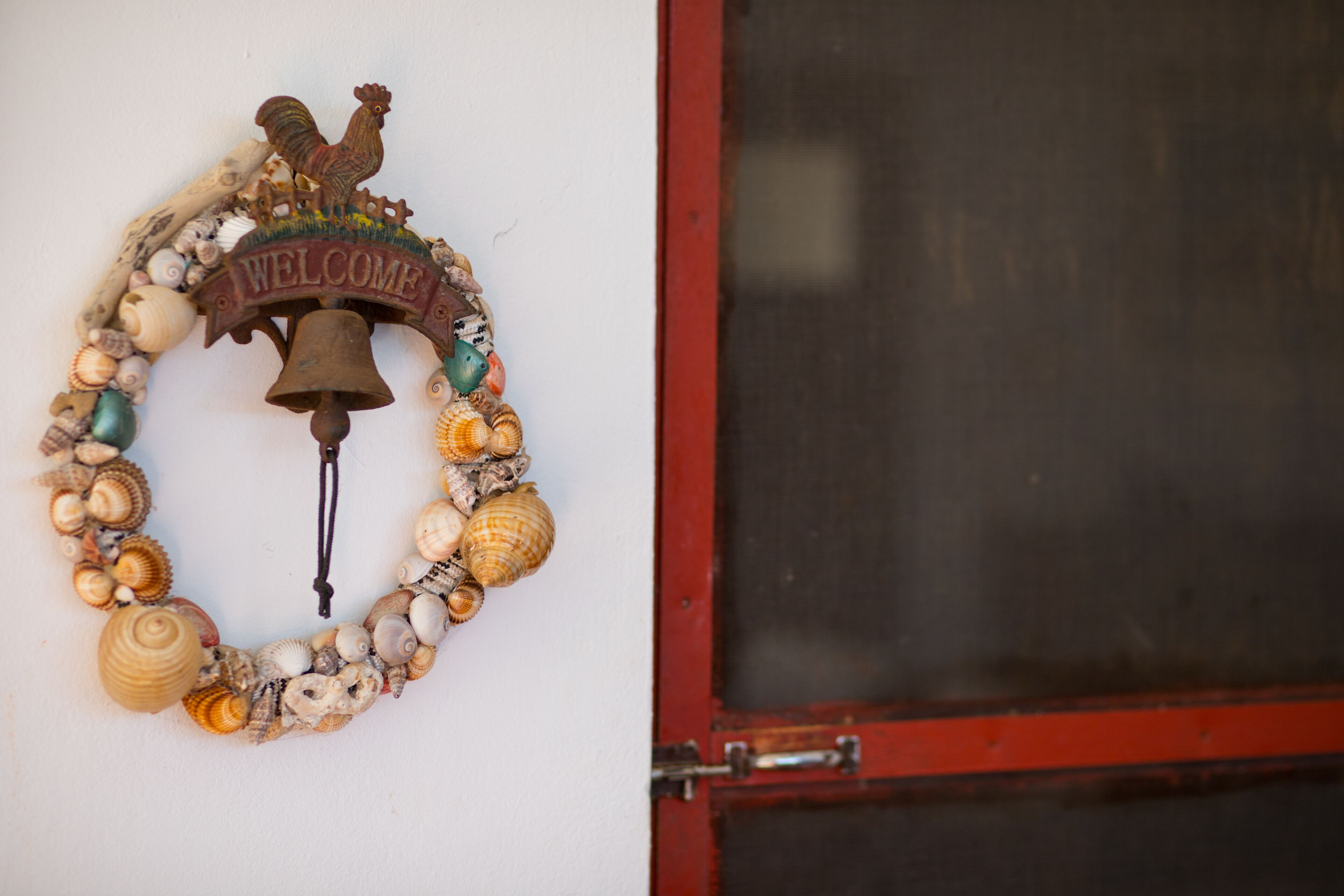 A welcome sign with a rooster and a bell, surrounded by seashells, mounted on a white wall next to a red door.