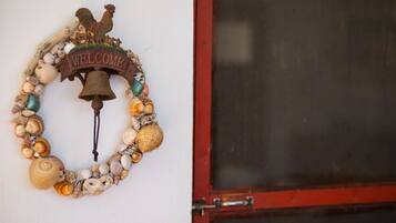 A welcome sign with a rooster and a bell, surrounded by seashells, mounted on a white wall next to a red door.
