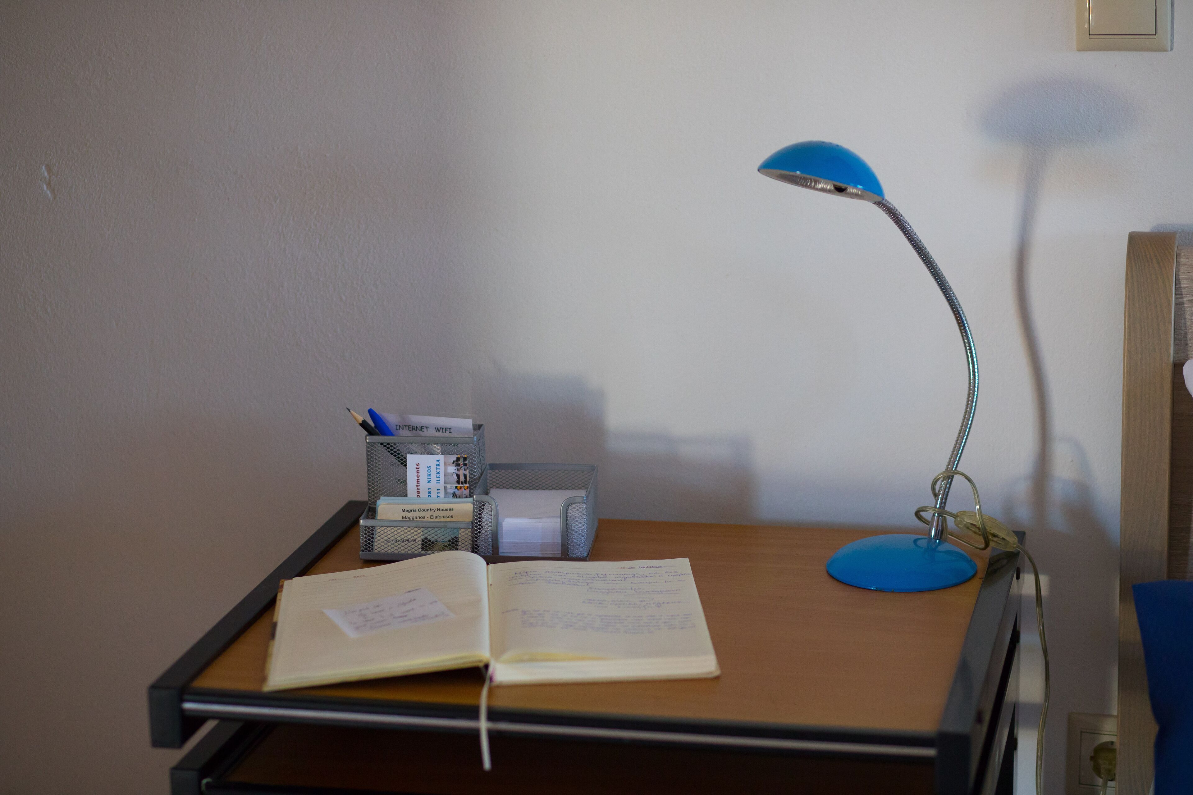 Ein Schreibtisch mit offenem Notizbuch, einer blauen Schreibtischlampe, einem Stifthalter und einer kleinen Schachtel mit Papier.
