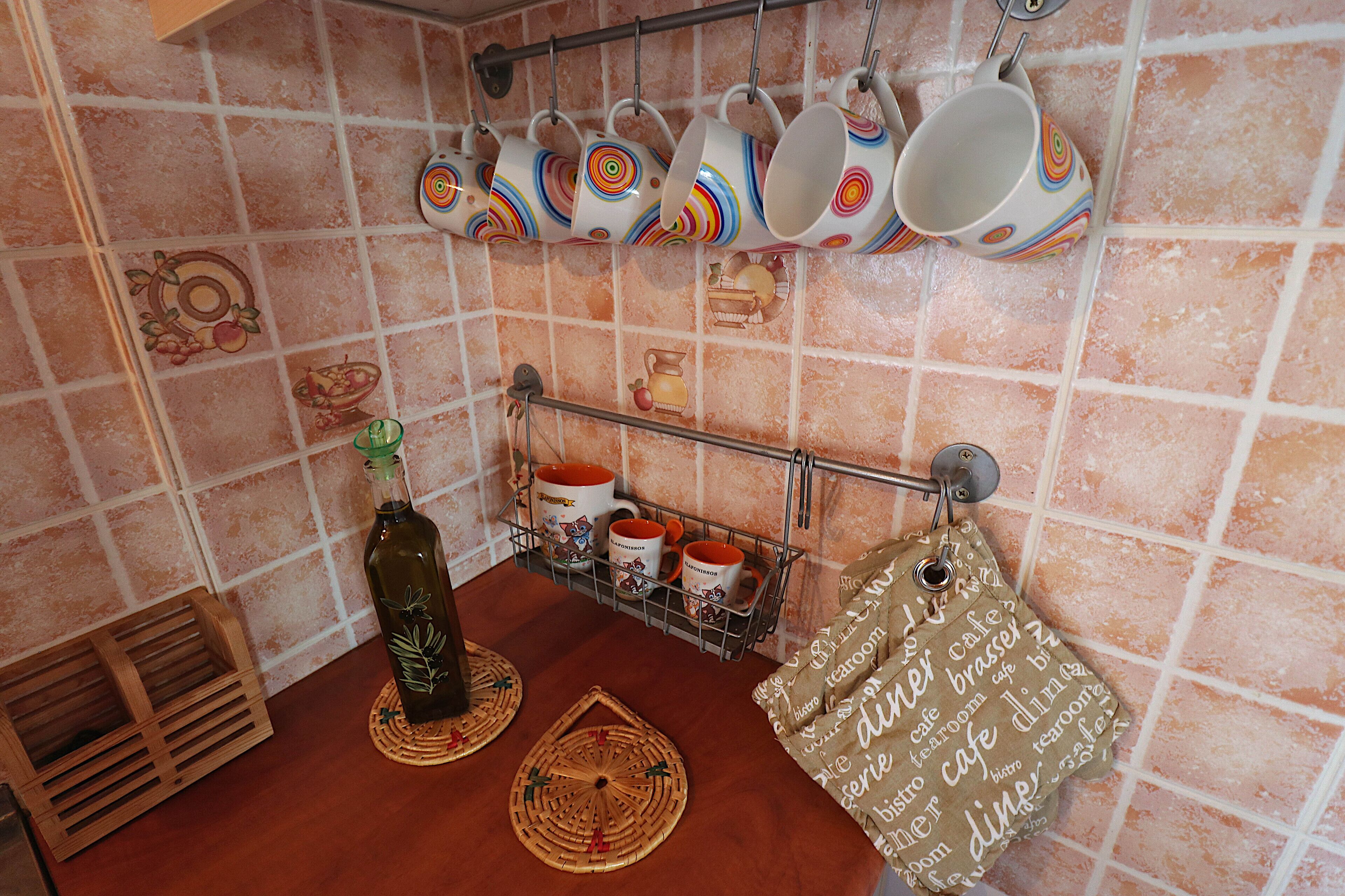 Una cocina con un colgador de tazas coloridas, una botella de aceite de oliva y un mantel tejido sobre la encimera.