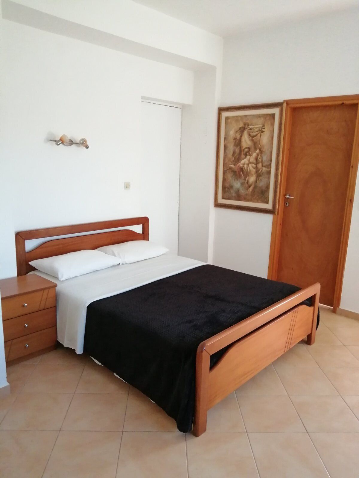 A bedroom with a wooden bed, a nightstand, a dresser, a door, and a painting on the wall.