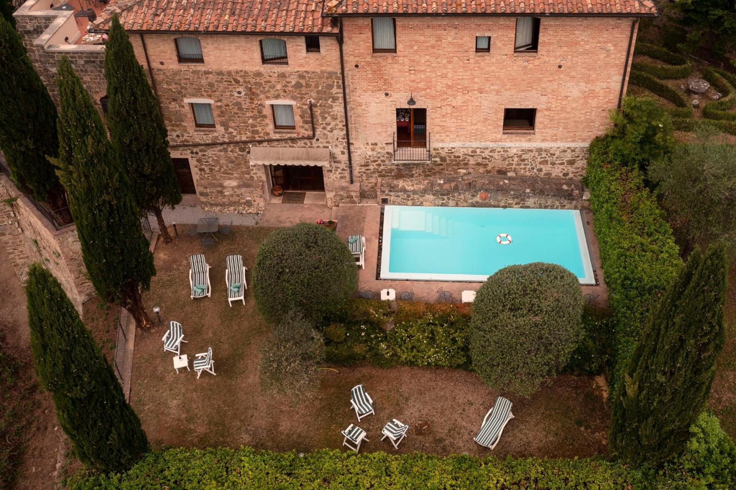 A large stone building with a swimming pool, surrounded by neatly trimmed hedges and several lounge chairs.