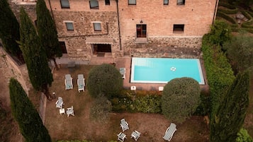 A large stone building with a swimming pool, surrounded by neatly trimmed hedges and several lounge chairs.