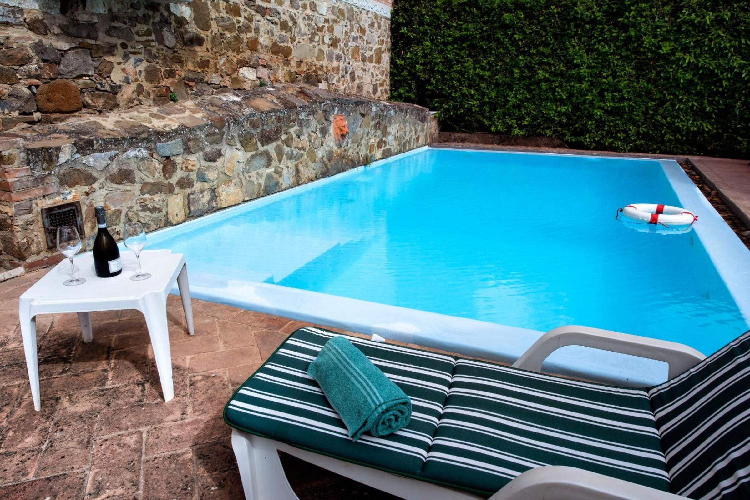 Une piscine bordée d’un mur en pierre et entourée d’une haie. On y trouve une chaise longue avec une serviette, ainsi qu’une petite table sur laquelle sont posés une bouteille et des verres.