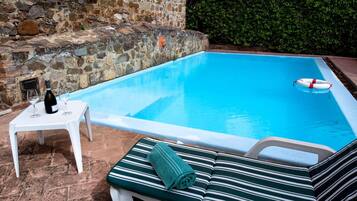 Une piscine bordée d’un mur en pierre et entourée d’une haie. On y trouve une chaise longue avec une serviette, ainsi qu’une petite table sur laquelle sont posés une bouteille et des verres.