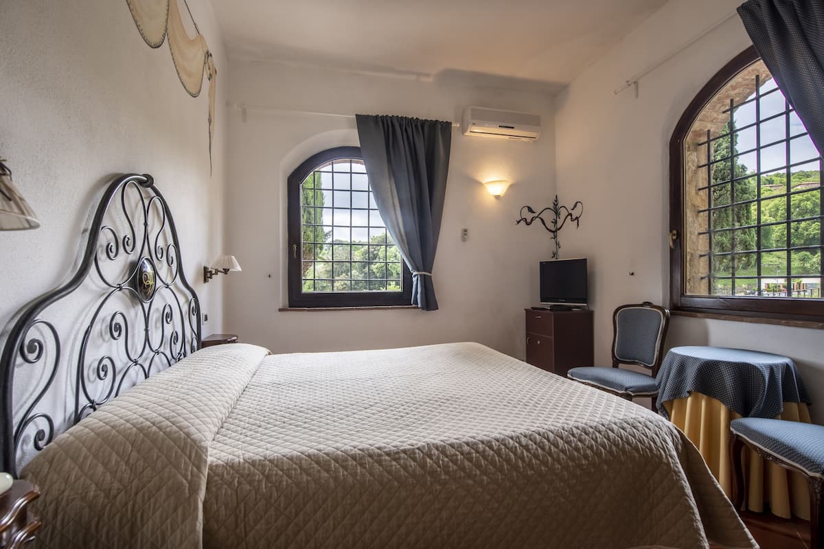 Ein Schlafzimmer mit einem Bett, einem Fernseher, einem Sessel, einem Tisch und einem fenster mit Vorhängen.