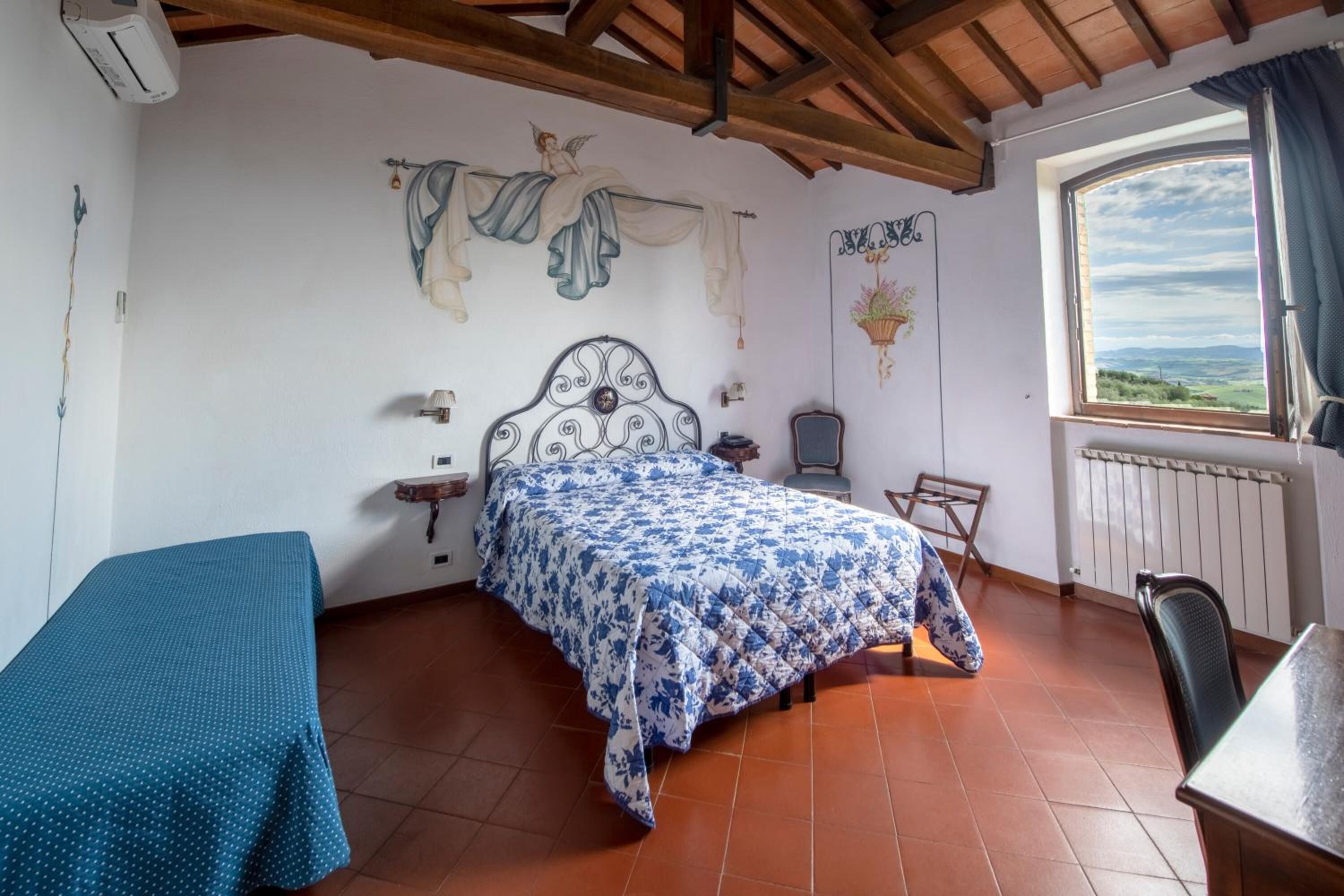 A bedroom with a bed, a chair, a table, a window with a view, and a wall with a hanging plant and artwork.