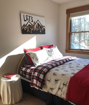 Quarto com cama, travesseiros, mesa de cabeceira, janela com vista para árvores e um letreiro na parede.