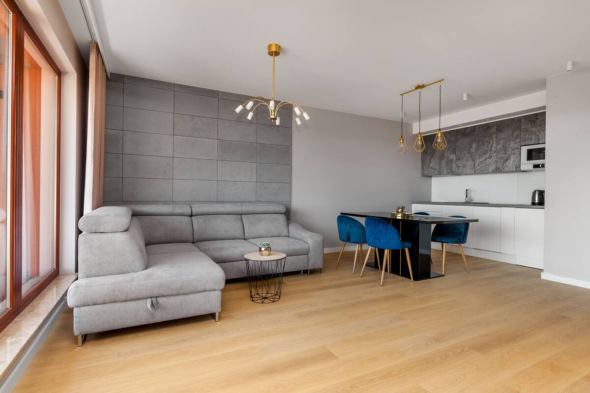 Ein modernes Wohnzimmer mit einem grauen Ecksofa, einem kleinen runden Tisch und einem Essbereich mit blauen Stühlen.