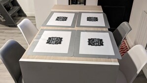 A modern dining table with four square placemats, each featuring a black and white logo. The table is surrounded by six white chairs with metal frames. - Guesthouse near Charles De Gaulle Airport (Thieux)