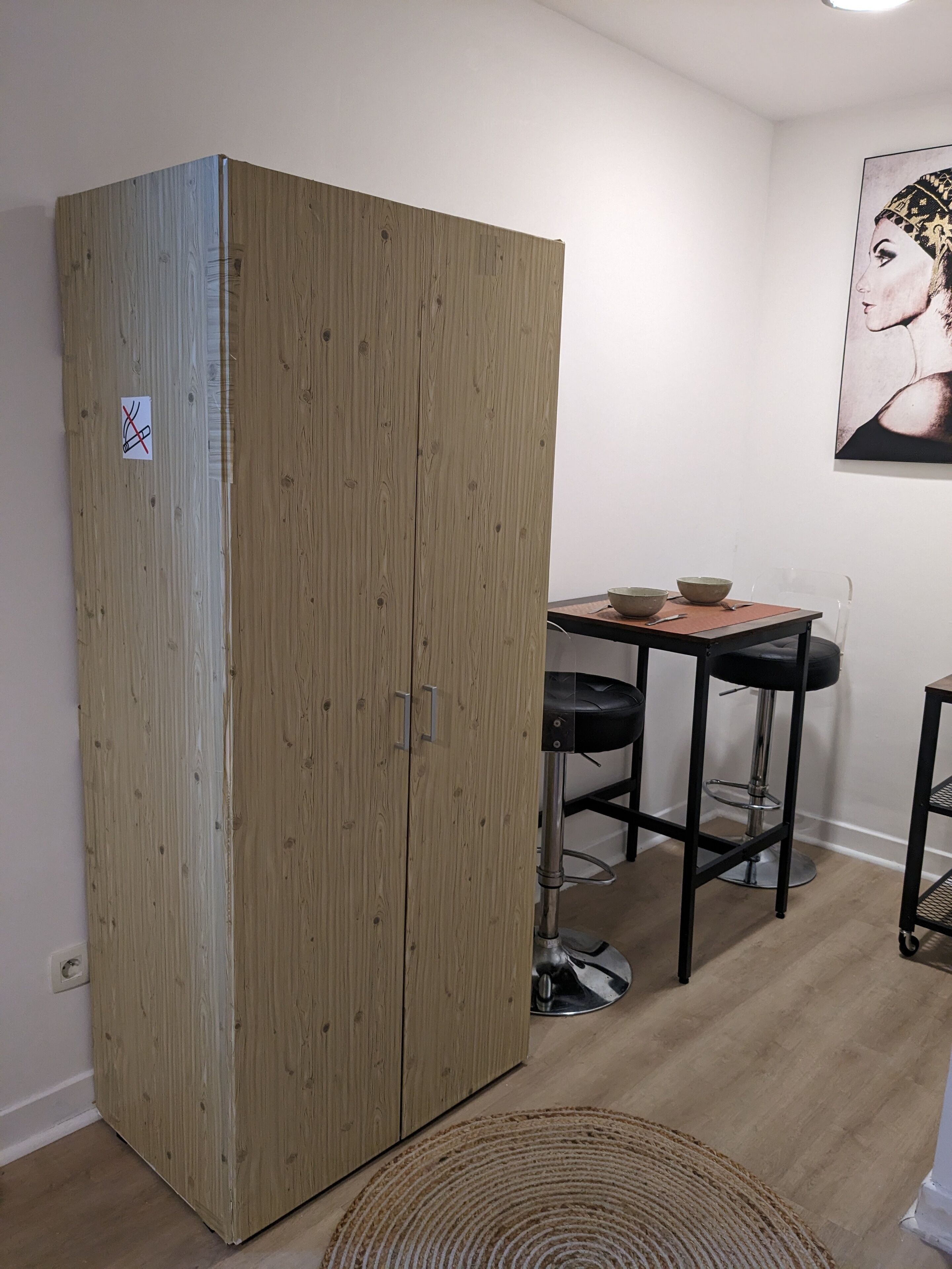 A room with a wooden wardrobe, a dining table with two chairs, and a bar stool.