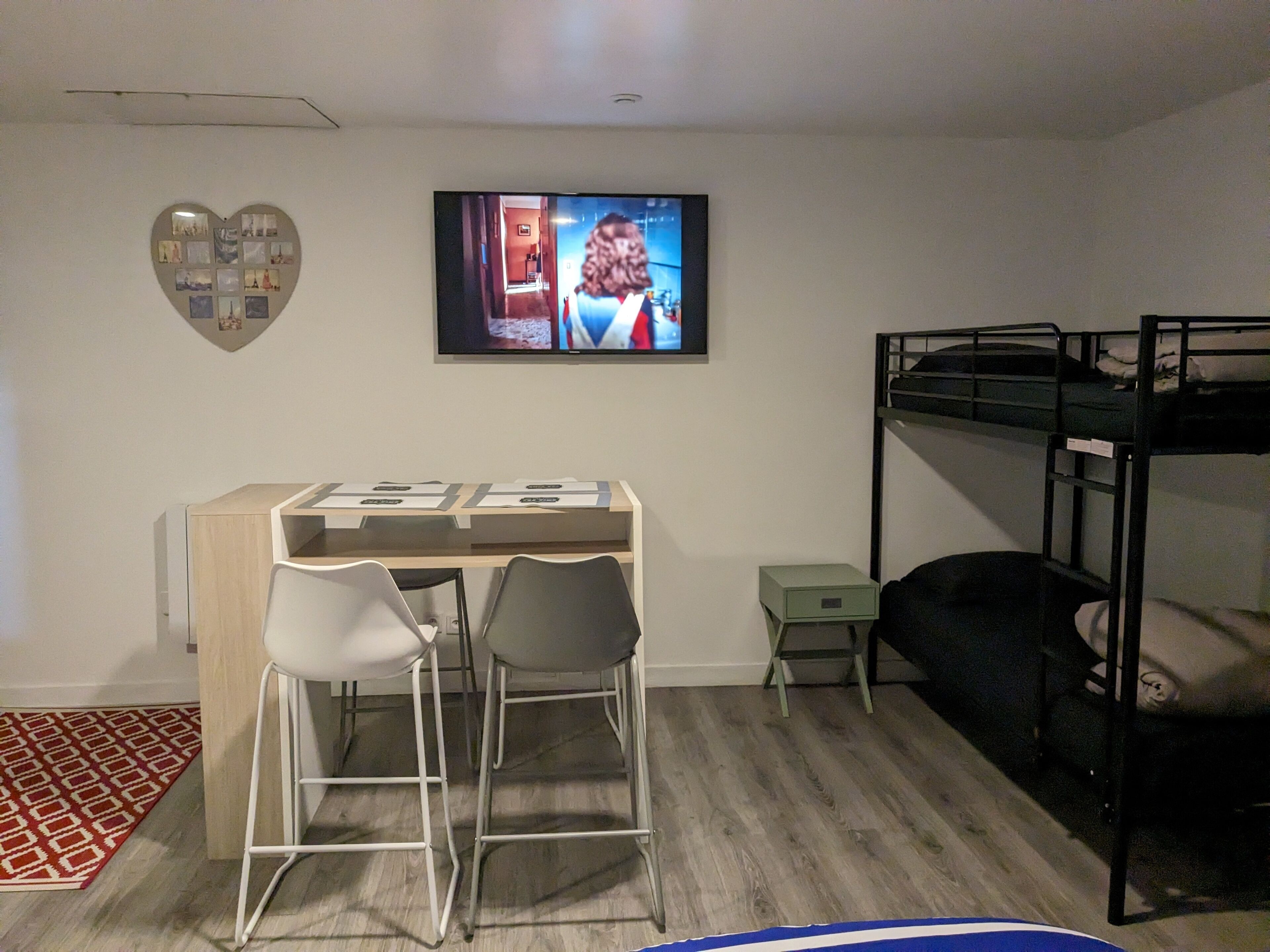 A bedroom with a bunk bed, a desk with two chairs, a television on the wall, and a heart-shaped photo collage.