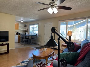 A living room with a ceiling fan, a television, a dining area with chairs, a kitchen in the background, and a large window with a view of the outside. - Thunder Mountain Place - Quiet, Comfortable Place Offering DISCOUNT ON TOURS! (Juneau)