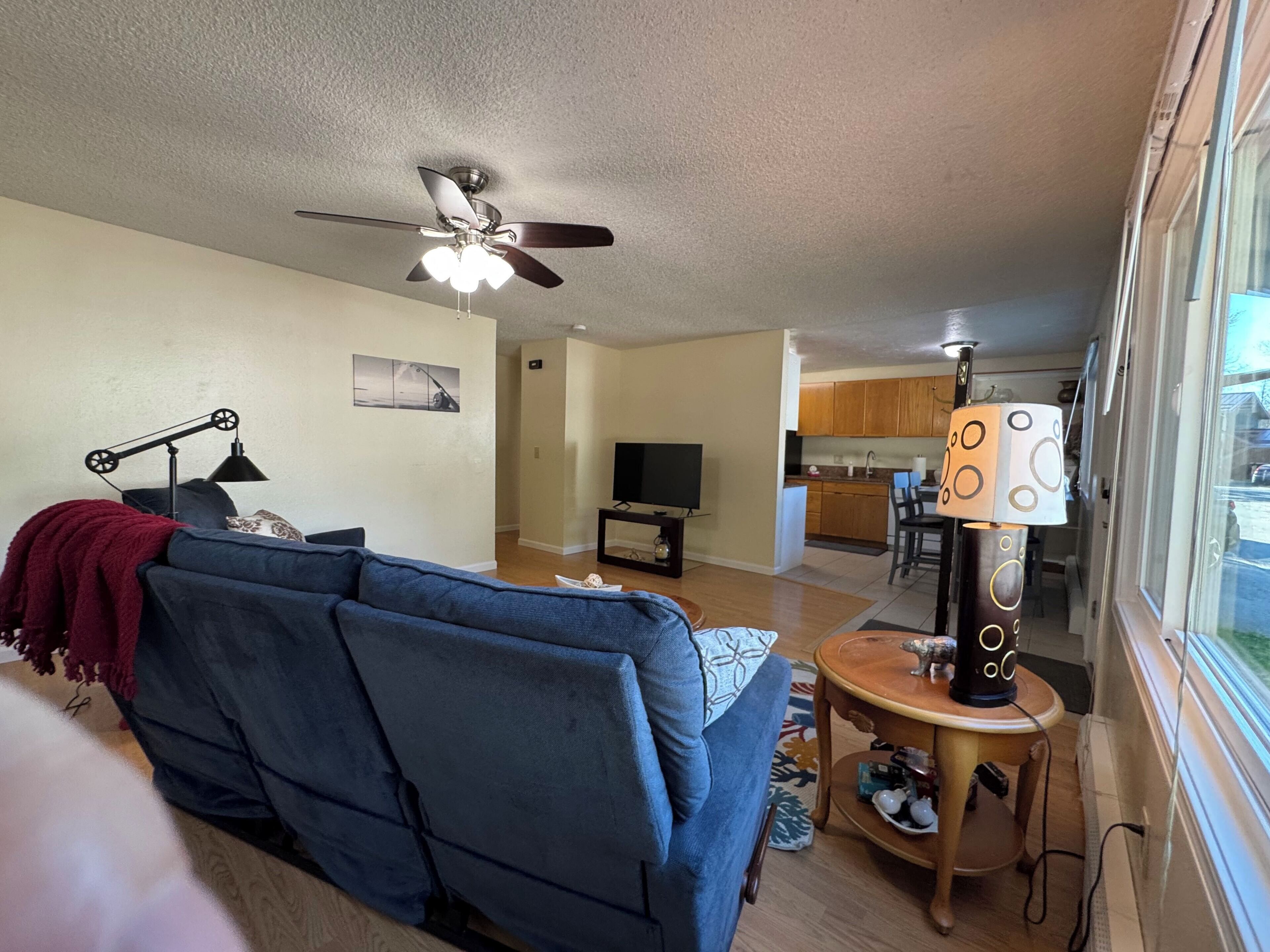 Ein Wohnzimmer mit blauem Sofa, Deckenventilator, Lampe und Blick auf eine Küche und Essbereich.