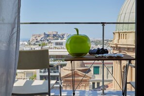 Varanda com uma maçã verde, binóculos e uma cadeira, com vista para um horizonte urbano com edifícios históricos.
