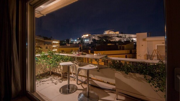 A balcony with two bar stools, a table, and potted plants. In the background, a building with illuminated windows is visible.