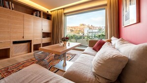 A modern living room with a sofa, coffee table, and bookshelf. A large window offers a view of the cityscape.
