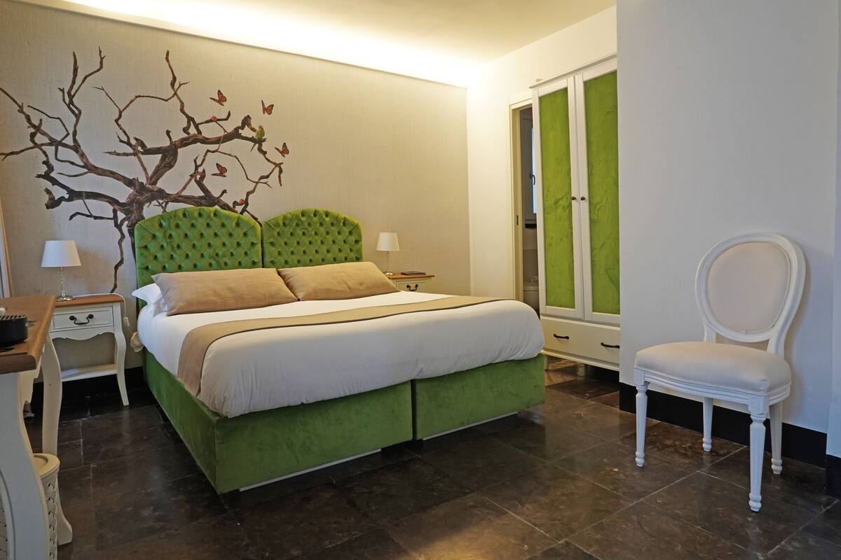 A bedroom with a green upholstered bed, a white chair, a nightstand with a lamp, and a wall mural.