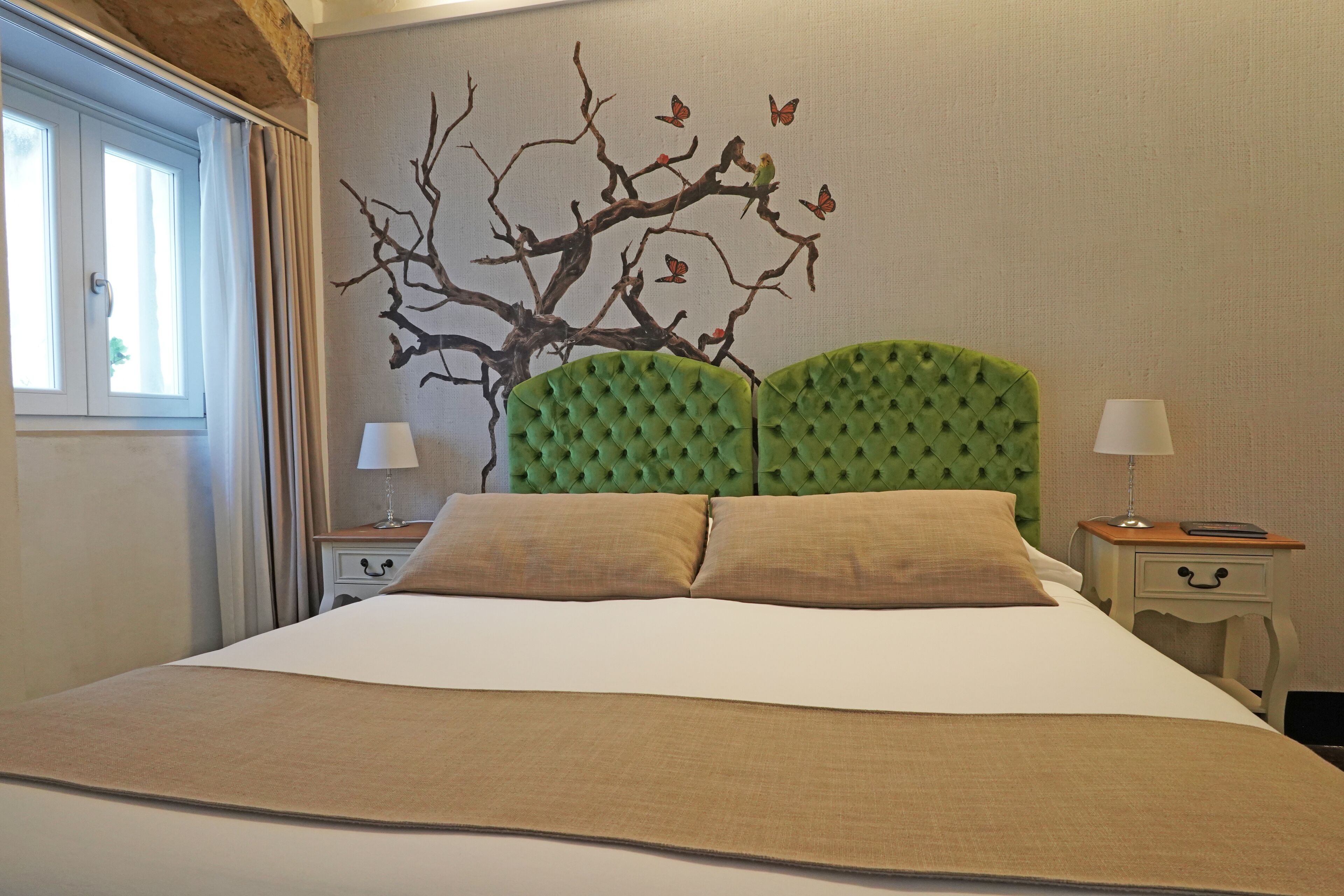 A neatly made bed with a green upholstered headboard, two bedside tables with lamps, and a wall mural of a tree with butterflies.