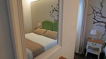 A bedroom with a large mirror reflecting the room, a green upholstered headboard, a bed with beige bedding, a nightstand with a lamp, and a wall with a branch design.