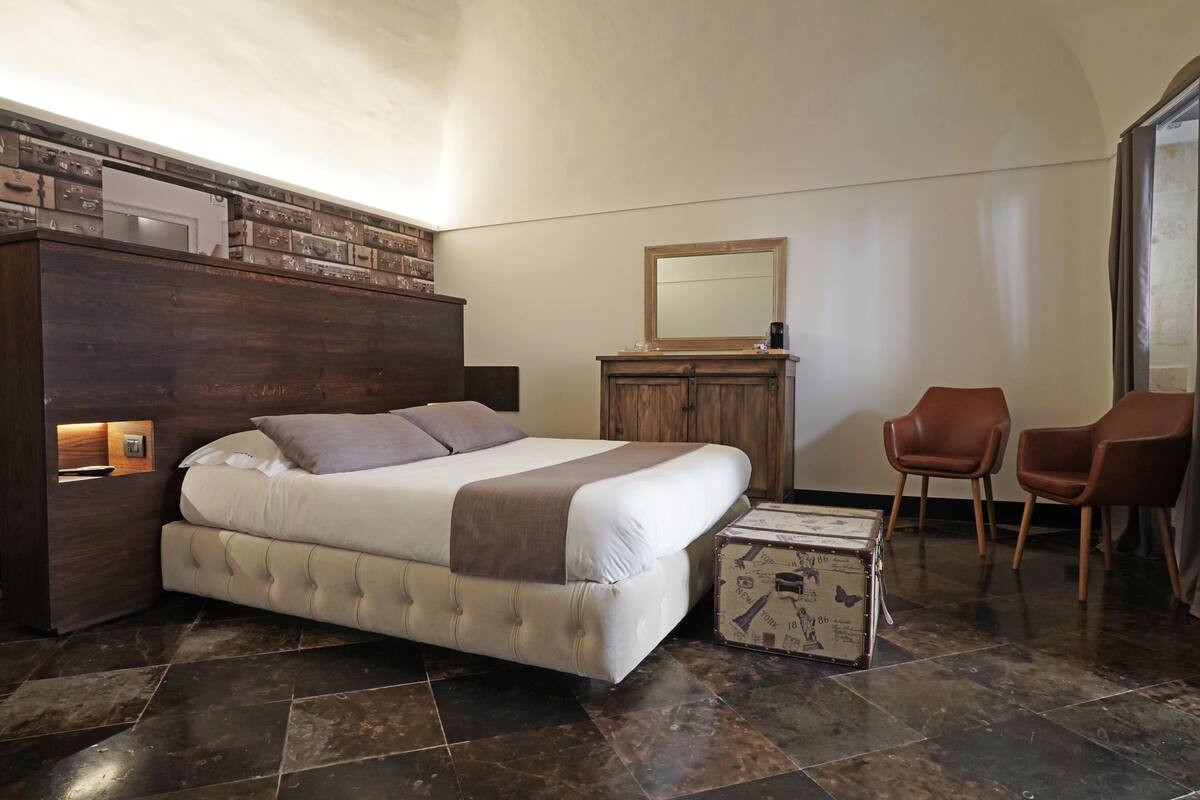 A bedroom with a large bed, a wooden dresser, two chairs, and a suitcase on the floor.