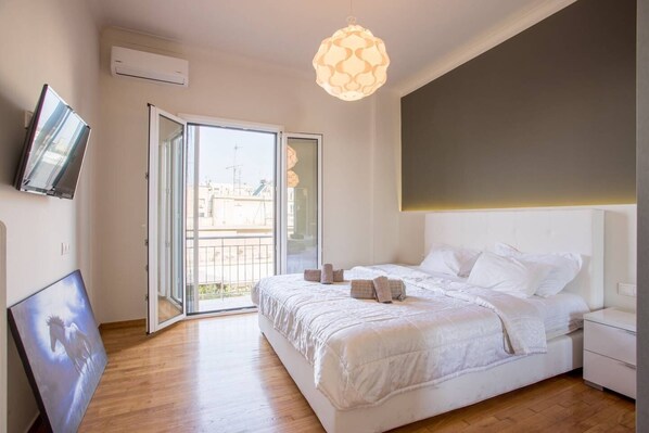 A modern bedroom with a large bed, a TV mounted on the wall, and a balcony with a view of the city.