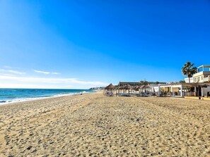 A beach with umbrellas and lounge chairs, a clear blue sky, and buildings in the background.