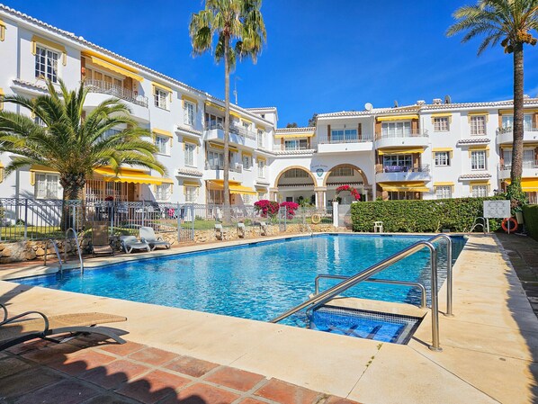 A swimming pool with a clear blue water, surrounded by a white building with yellow accents and multiple balconies.