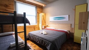 Una habitación con literas, un armario de madera, una cama con manta roja y un estante fijado a la pared.