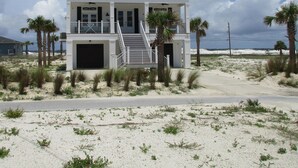 Ein zweistöckiges Strandhaus mit einem Balkon, einer Treppe, die zum Eingang führt, und einem Sandstrand im Hintergrund.