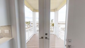 Blick vom Balkon mit Holzboden und weißen Geländern auf einen Strand und Palmen.