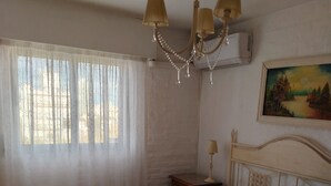 A hotel room with a large bed, a nightstand, a chandelier, a painting on the wall, and a window with curtains.