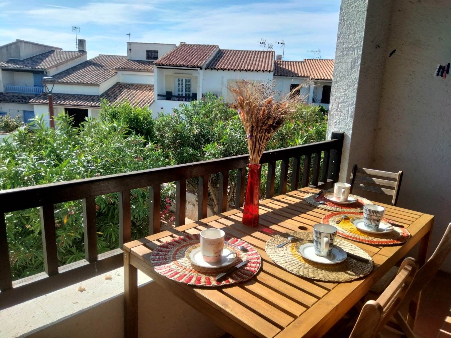 A wooden table set with a vase, plates, and cups on a balcony with a view of houses and greenery.