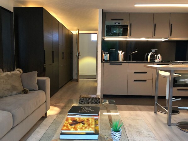 A modern living room with a sofa, a coffee table with books, and a kitchen area with cabinets and appliances.