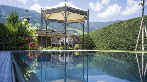 Une piscine aux eaux cristallines, agrémentée d’un belvédère et de chaises, entourée de verdure et avec des montagnes en toile de fond.