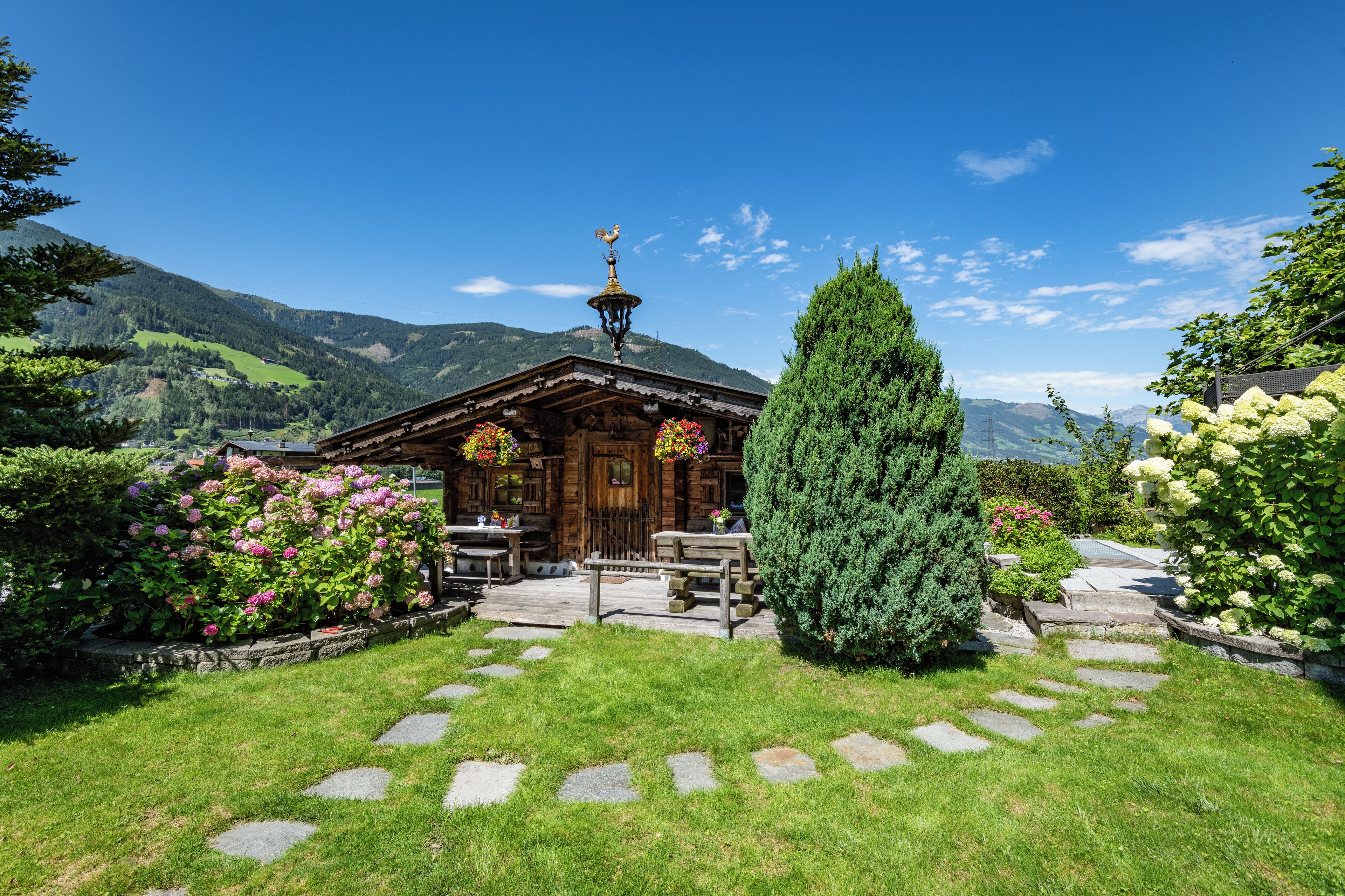 Un chalet en bois, avec un chemin de pierre menant à l’entrée, entouré d’une végétation luxuriante et dominé par un paysage montagneux.