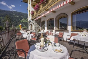 An outdoor dining area with tables and chairs, a mountain view, and a building with stone walls and flower boxes. - Hotel Riedl (Stumm)