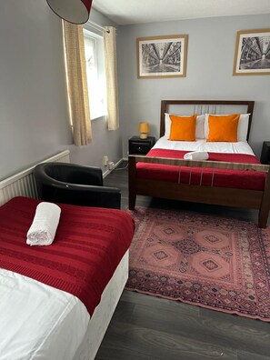 A hotel room with two beds, a red and white bedspread, a wooden headboard, a nightstand with a lamp, a patterned rug, and two framed pictures on the wall. - Poynters House - Huku Kwetu Luton (Luton)