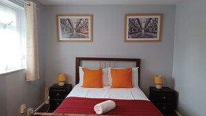 A neatly arranged hotel room with a large bed, two nightstands, a window with curtains, and two framed pictures on the wall. - Poynters House - Huku Kwetu Luton (Luton)