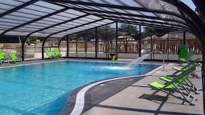 A covered swimming pool with clear blue water, surrounded by green lounge chairs and a glass enclosure. - Pluvier argenté (Quiberon)