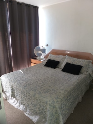 A bed with a floral bedspread, two black pillows, a wooden nightstand with a fan, and a window with curtains. - Vadigi (Santiago)