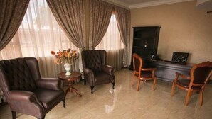 Un salón espacioso con un sofá de cuero grande, un sillón más pequeño y una silla de madera. Una mesita con un florero está en el centro. La habitación tiene ventanas grandes con cortinas y una chimenea.