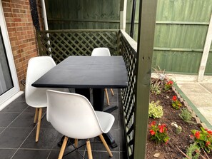 Eine Terrasse mit einem schwarzen Tisch und weißen Stühlen, umgeben von einem Holzzaun und einem Garten mit roten Blumen.